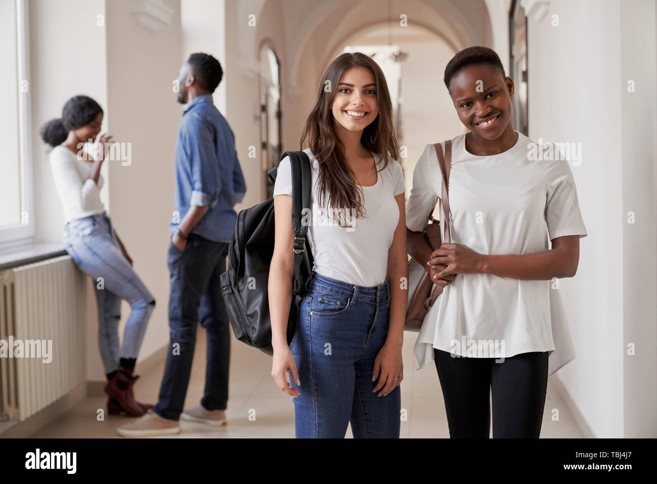 Piuttosto africano e gli studenti caucasici ragazze in piedi sul corridoio di università e sorridente alla fotocamera. Le donne mantenendo gli amici, studiare in università internazionale insieme. Foto Stock