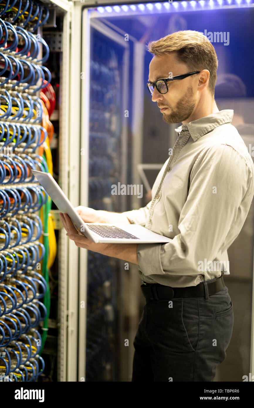 Grave concentrata qualificato programmatore barbuto in bicchieri in piedi dal cabinet per server e utilizzando computer portatile mentre esaminando server Foto Stock