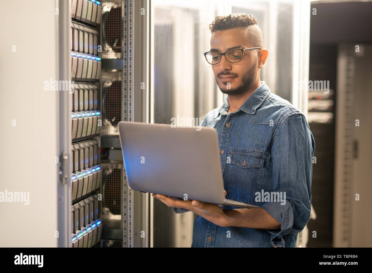 Grave giovane esperto Arabian specialista IT con la barba in piedi da moderni server e utilizzando computer portatile durante il monitoraggio di processi di rete Foto Stock