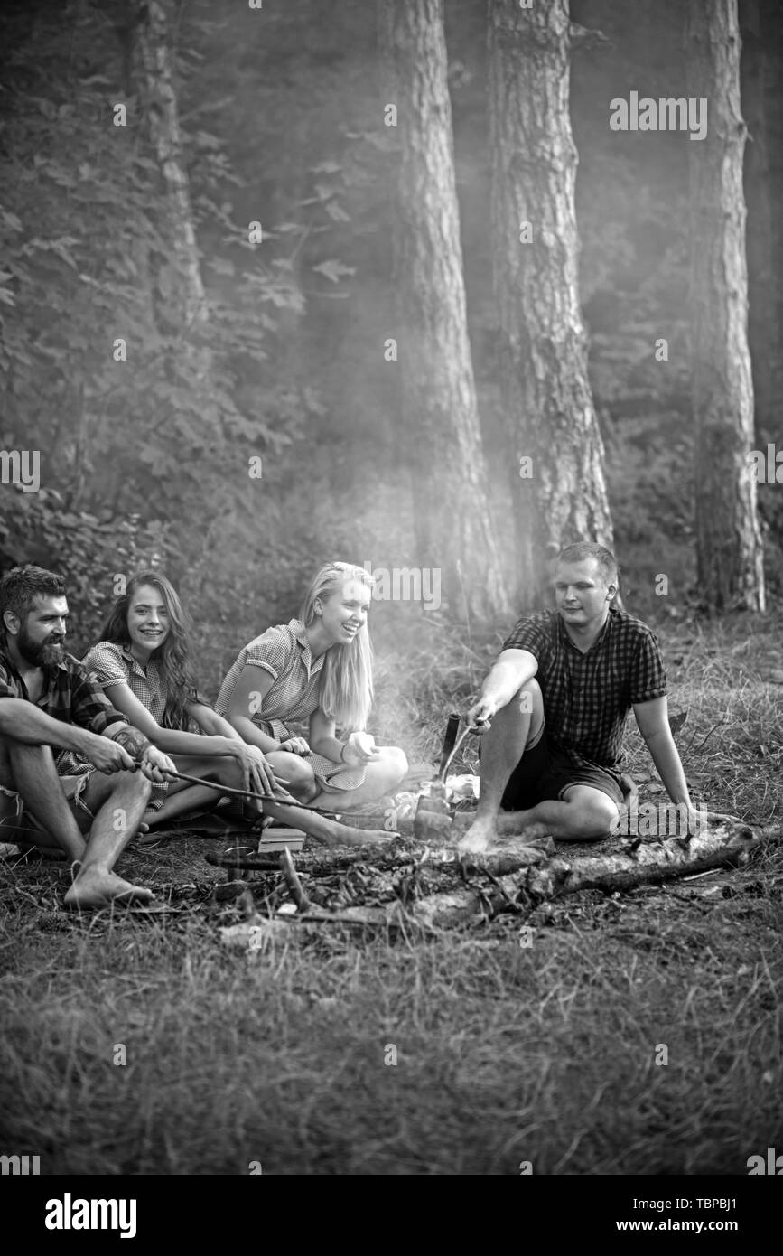 Happy amici seduti intorno al falò in serata, amicizia e il concetto di tempo libero. Giovani coppie sorridente avente picnic nei boschi. Uomo Barbuto Foto Stock