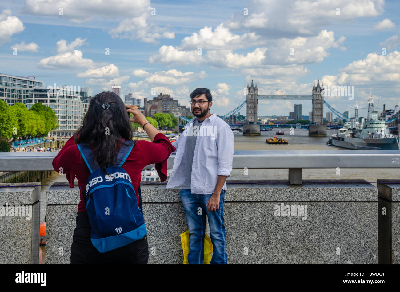 Una signora prende una fotografia del suo compagno sul Ponte di Londra con il fiume Tamigi, HMS Belfast e Tower Bridge in background. Foto Stock