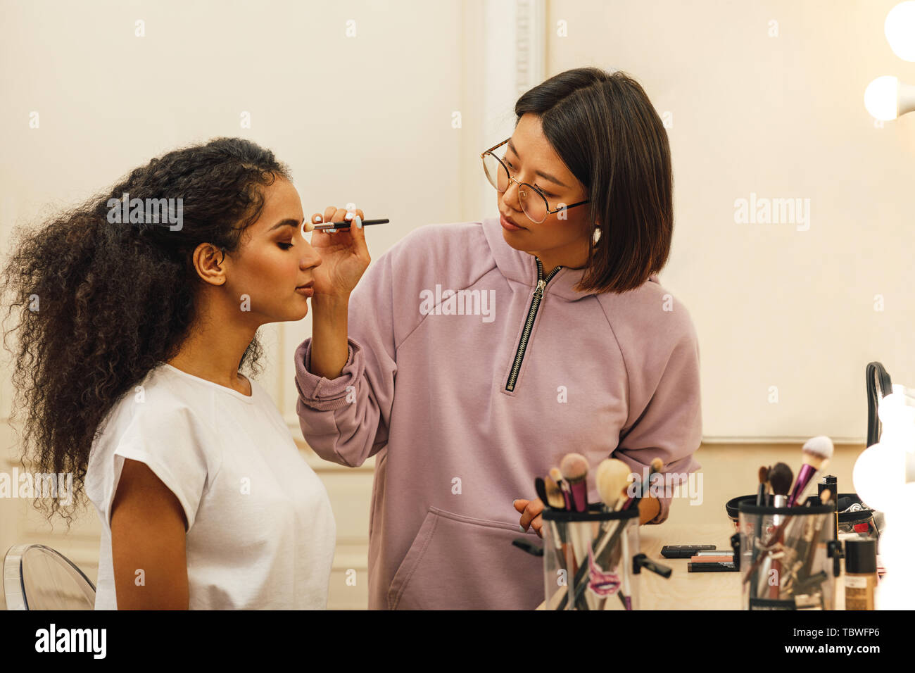 Vista laterale di un modello di seduta in una dressing room mentre truccatore applicando makeover sul suo occhio Foto Stock