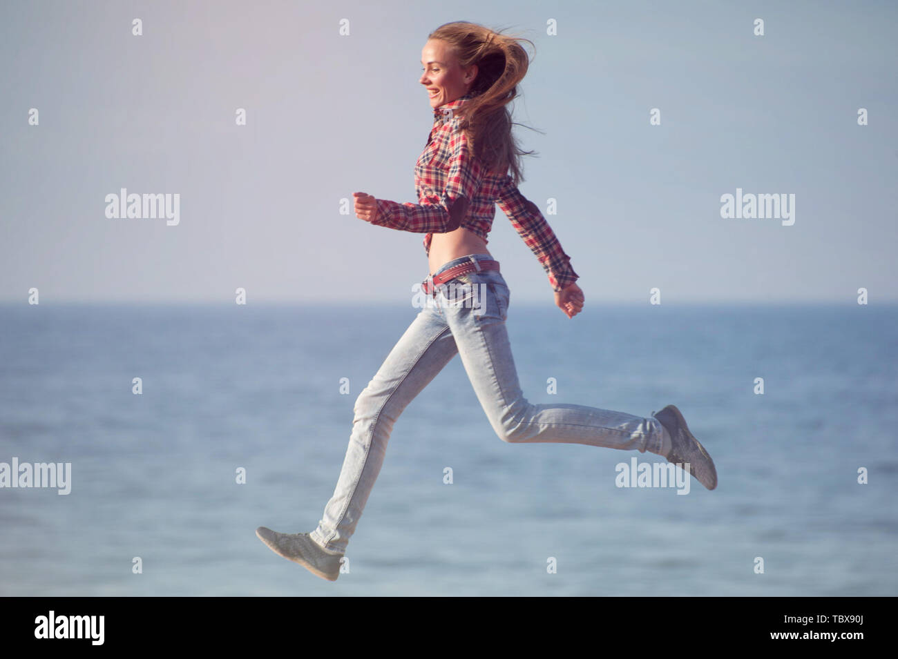 Felice active giovane femmina in plaid shirt che corre lungo la costa dell'oceano in estate tramonto Foto Stock