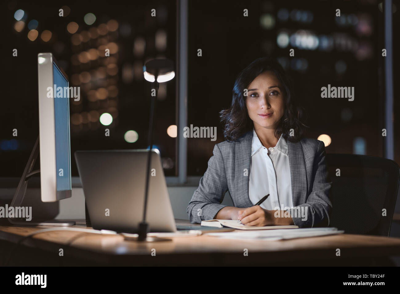 Imprenditrice sorridente seduto alla sua scrivania in un ufficio scuro Foto Stock