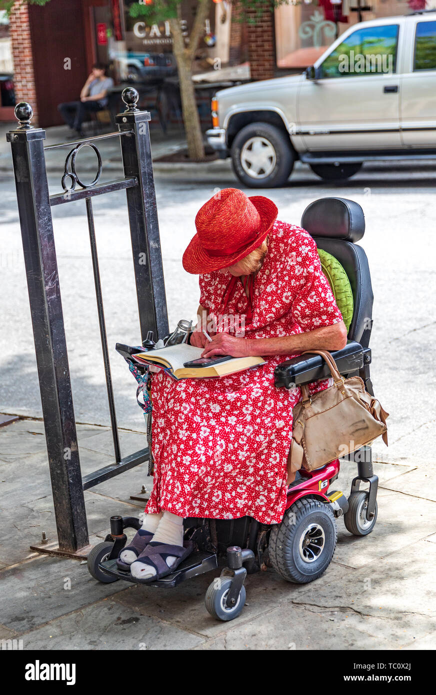 ASHEVILLE, NC, Stati Uniti d'America-5/31/19: a portatori di handicap, donna anziana in rosso brillante abito di stampa e Red Hat, siede in sedia a rotelle e la lettura di un libro e di fumare. Foto Stock