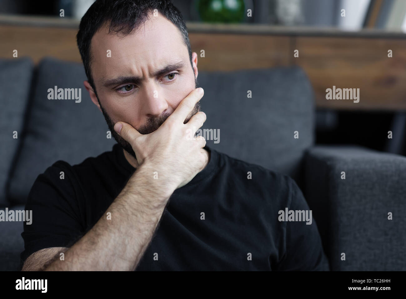 Grave man in black t-shirt tenendo la mano sulla bocca mentre seduti a casa Foto Stock