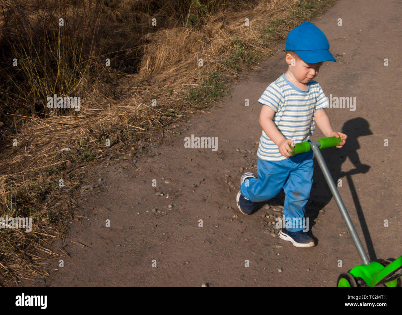 Inizio della primavera, un bimbo di tre anni in pantaloni blu con uno scooter verde sulla strada di un paese. Foto Stock