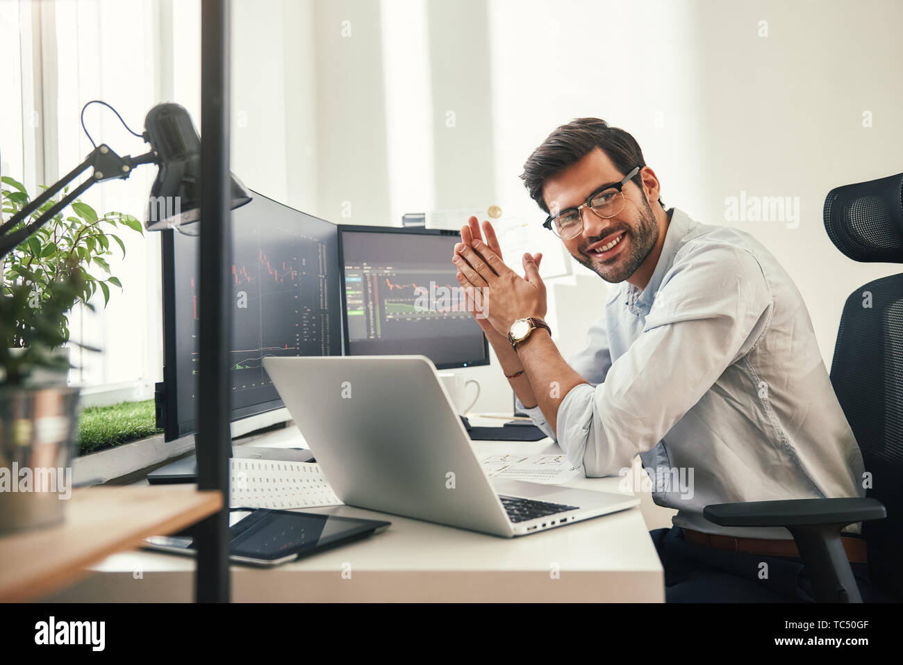 Giovani e di successo. Felice barbuto trader formale indossare occhiali e guardando la telecamera e sorridere mentre seduto nel suo ufficio moderno. Stock broker. Mercato Forex. Concetto di commercio Foto Stock