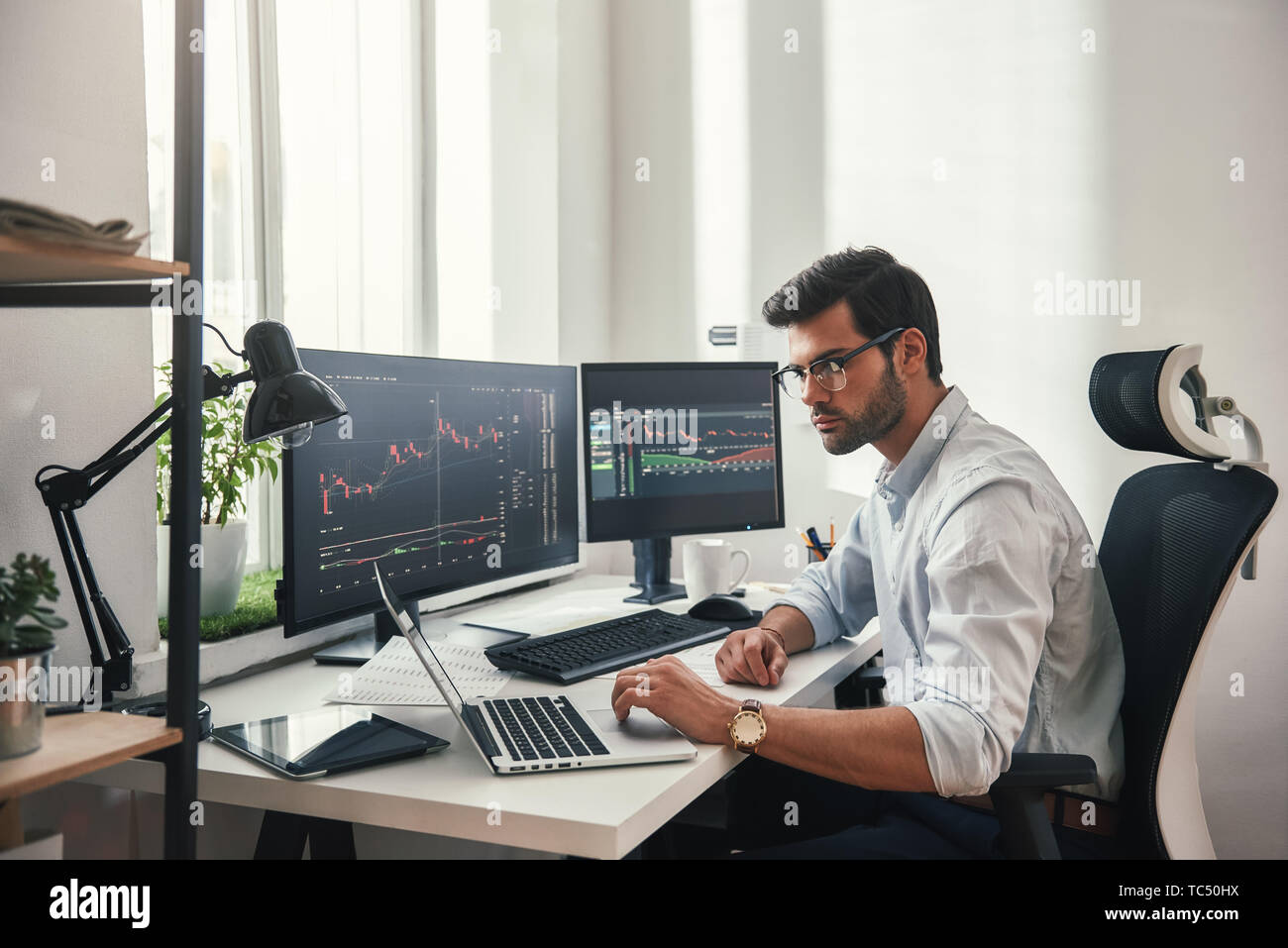 Intensa giornata di lavoro. Giovani barbuto trader in occhiali a lavorare  con il computer portatile mentre è seduto nel suo ufficio moderno davanti  allo schermo del computer con i grafici di trading.