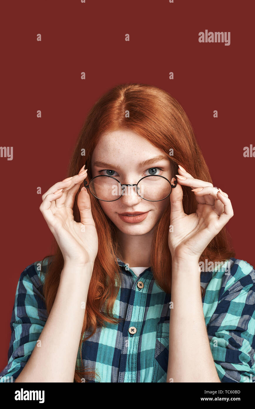 Close up della cute redhead ragazza con i capelli lunghi toccando specs e guardando la telecamera indossando camicia a scacchi isolate su sfondo rosso. Colpo verticale Foto Stock
