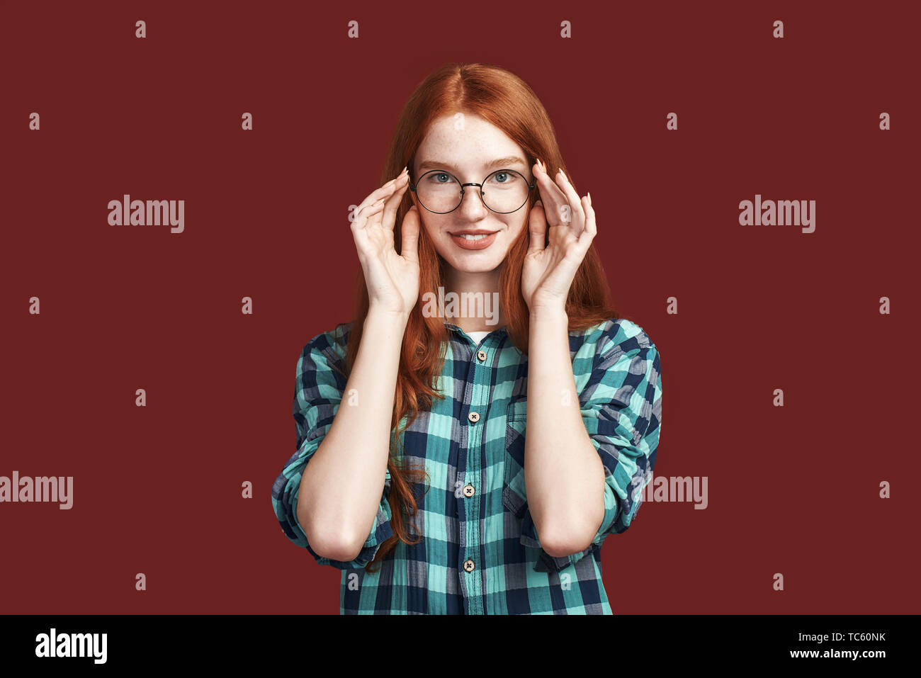 Close up della cute redhead ragazza con i capelli lunghi toccando specs e guardando la telecamera indossando camicia a scacchi isolate su sfondo rosso. Inquadratura orizzontale Foto Stock