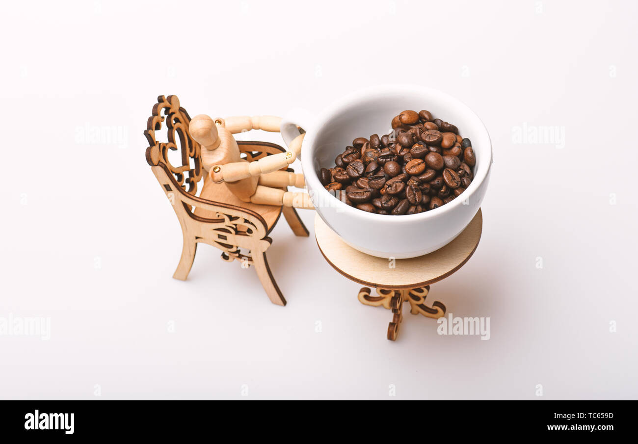 Pausa caffè concetto. Mug freschi chicchi di caffè tostati guarda gigante sul piccolo tavolo. Tazza di chicchi di caffè piccolo tavolo decorativo con sedia e il modello in legno di umana isolata su bianco. Foto Stock