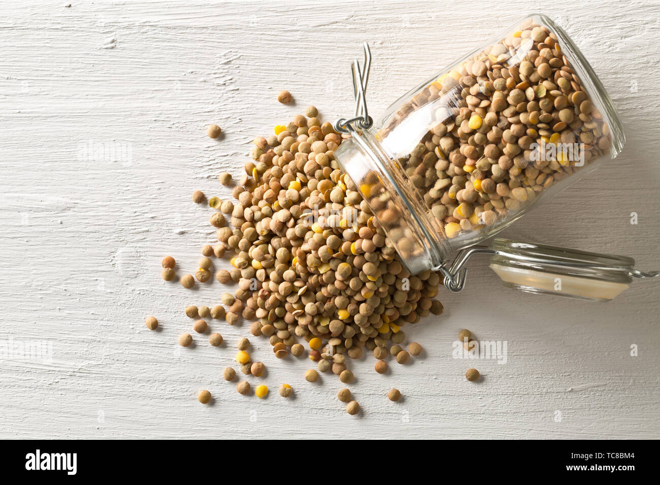 Raw, secco, non cotte lenticchia marrone legumi in vetro vaso di storage su legno bianco sullo sfondo della tabella con il fuoco selettivo flat top laici vista da sopra Foto Stock