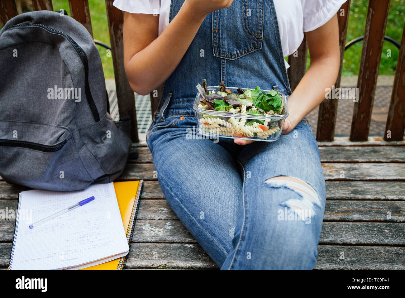 Sezione mediana verticale di uno studente ragazza seduta in un banco mentre mangiare sano con insalata di pasta in un bicchiere scatola di pranzo Foto Stock