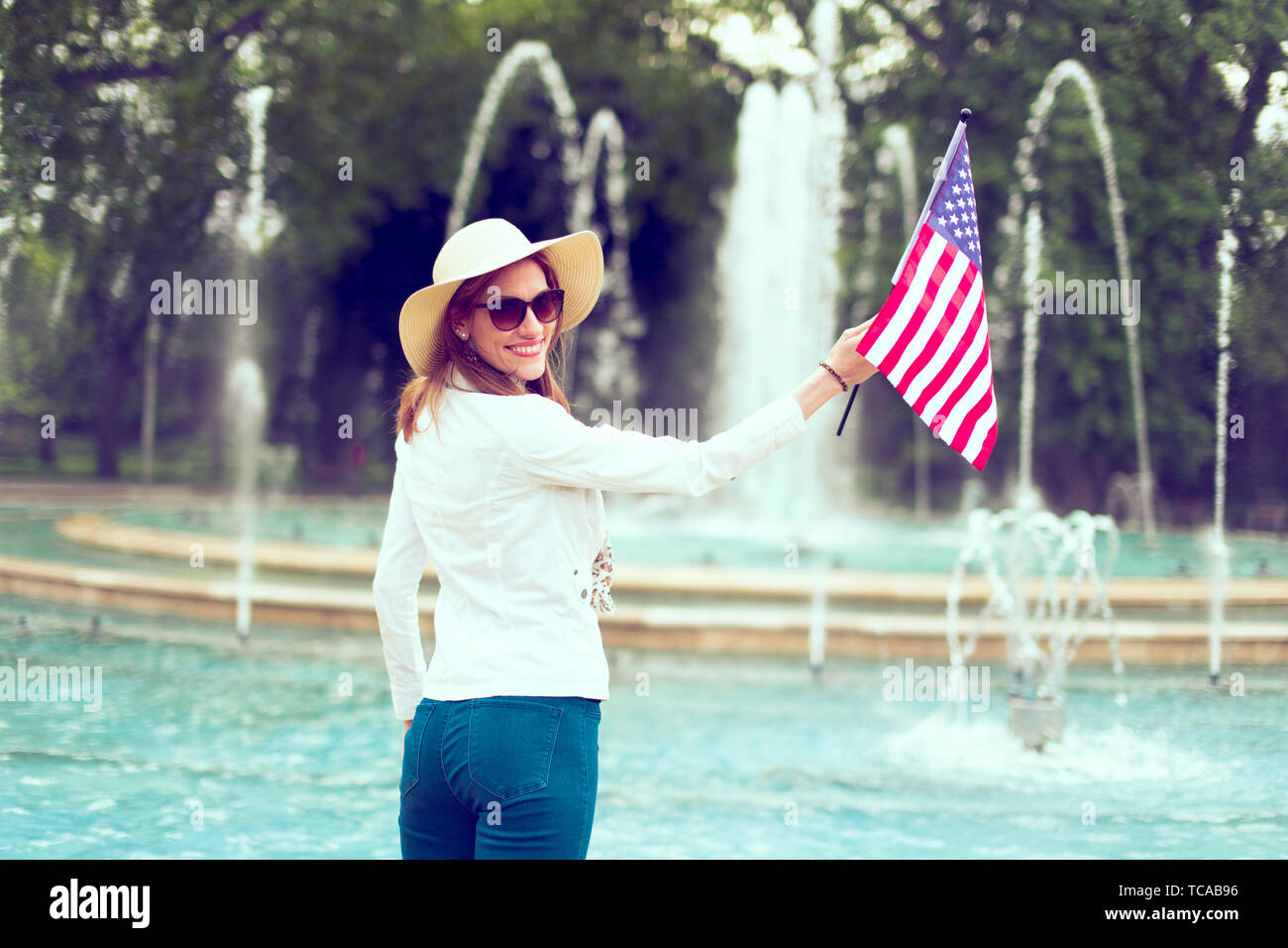 Patriot woman in hat holding bandiera degli Stati Uniti nel parco vista posteriore, guardando indietro, sorriso toothy, giorno di indipendenza, 4 luglio, in stile vintage Foto Stock