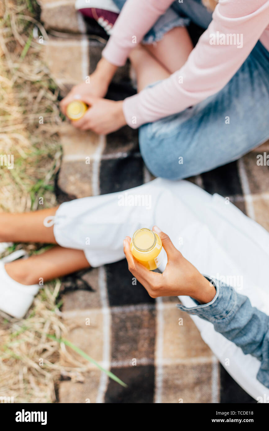 Vista ritagliata di due ragazze azienda bottiglie di succo di arancia seduti su plaid coperta sul terreno Foto Stock