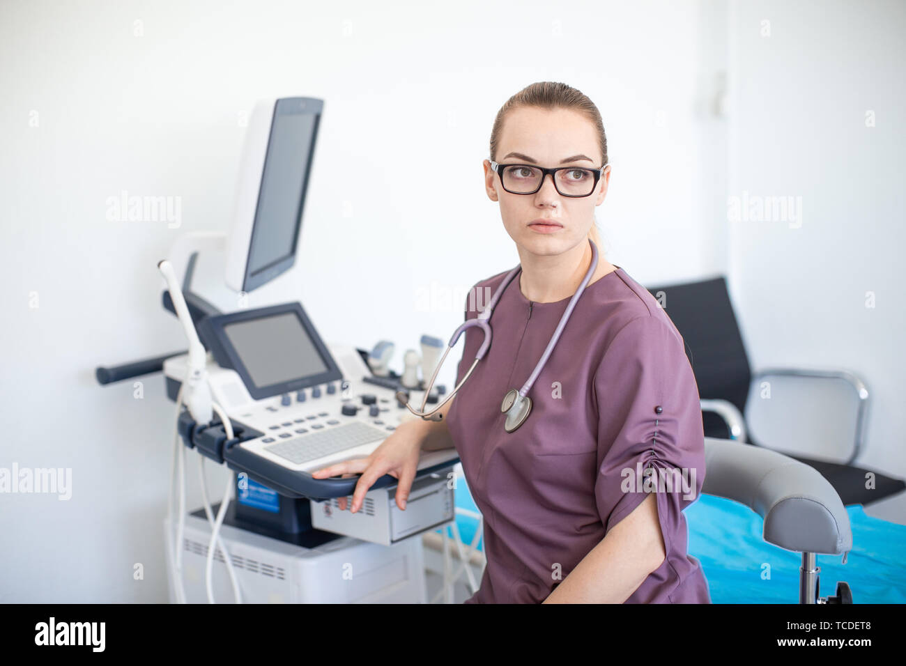 Giovane donna bionda medico in puprle uniforme con bicchieri e uno stetoscopio sul suo collo in clinica vicino a macchina ad ultrasuoni Foto Stock