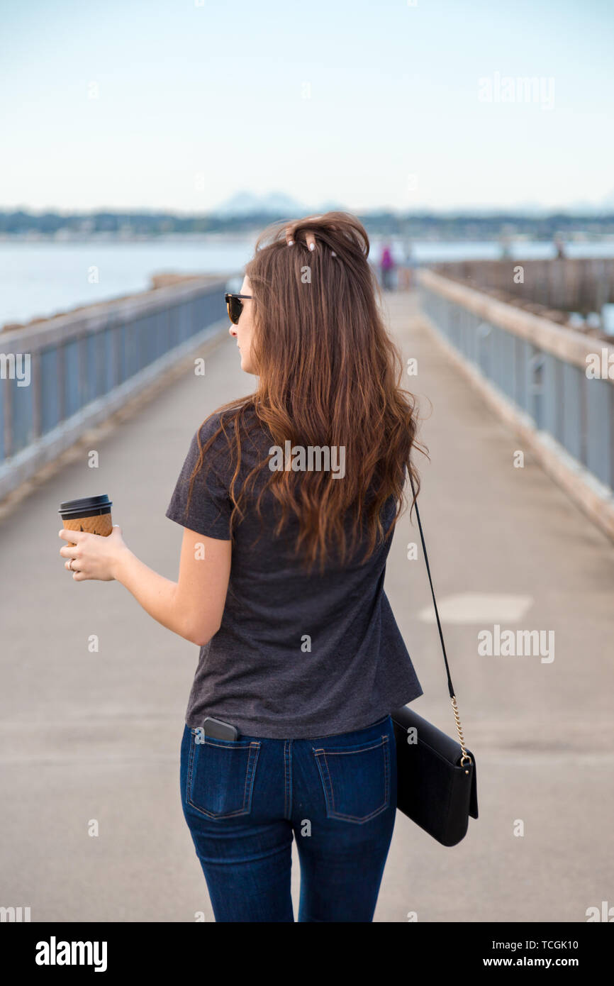 Una bella bruna giovane donna che indossa gli occhiali da sole con un caffè in un'Oceanside lungomare godendo la vista dell'oceano. Foto Stock