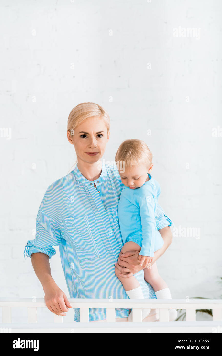 Attraente madre tenendo in armi toddler figlio e di stare vicino a culla a casa Foto Stock