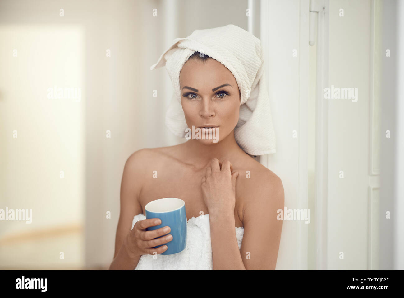 Bella donna di mezza età avvolto nelle fresche e pulite asciugamani bianchi attorno alla sua testa e corpo appoggiato contro una parete interna con una tazza di bevanda in un cost Foto Stock