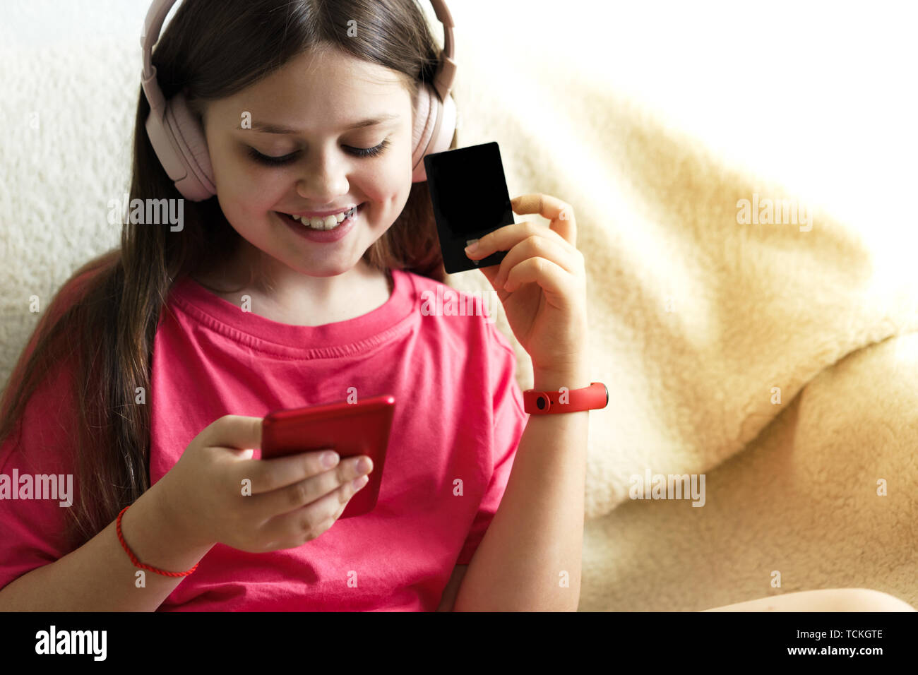 Allegro ragazza in rosa cuffie e una T-shirt rossa si siede con un telefono in mano e una carta di credito. Il concetto di gadget e i bambini di shopping onli Foto Stock