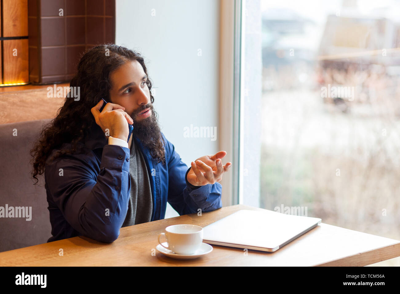 Ritratto di grave intelligenza bello giovane adulto uomo libero professionista in uno stile informale in seduta cafe con laptop, parlando al telefono, guardando lontano, Foto Stock