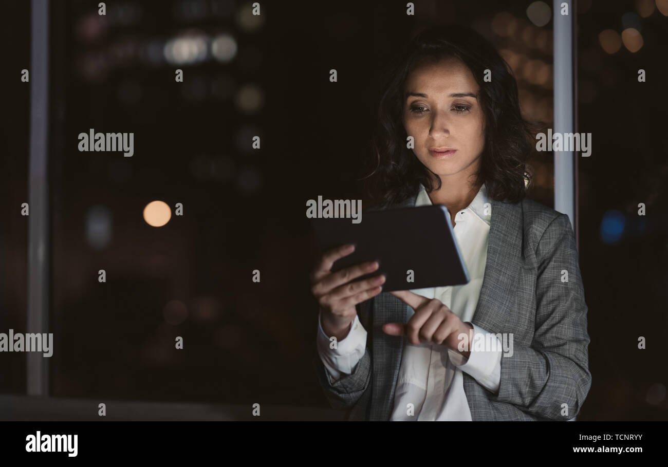 Focalizzato giovane imprenditrice in piedi da solo in un ufficio a lavorare con una tavoletta digitale a tarda notte con le luci della città in background Foto Stock