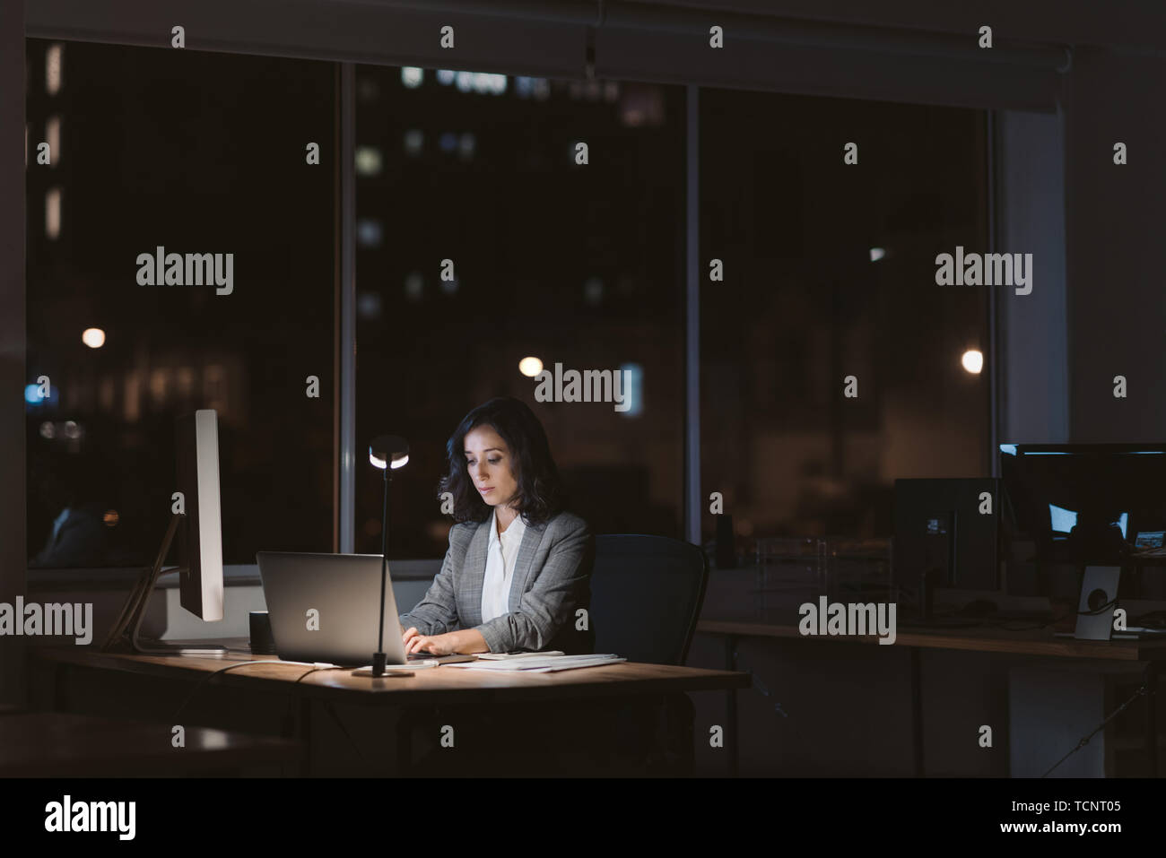 Giovane imprenditrice usando un computer portatile alla sua scrivania in un ufficio mentre si lavora fino a tardi la notte con le luci della città incandescente in background Foto Stock