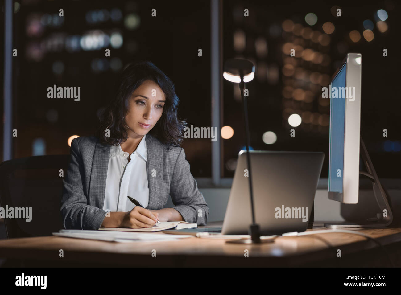 Giovane imprenditrice seduto alla sua scrivania utilizzando un computer portatile e la scrittura di note durante il lavoro straordinario in un ufficio a tarda sera Foto Stock