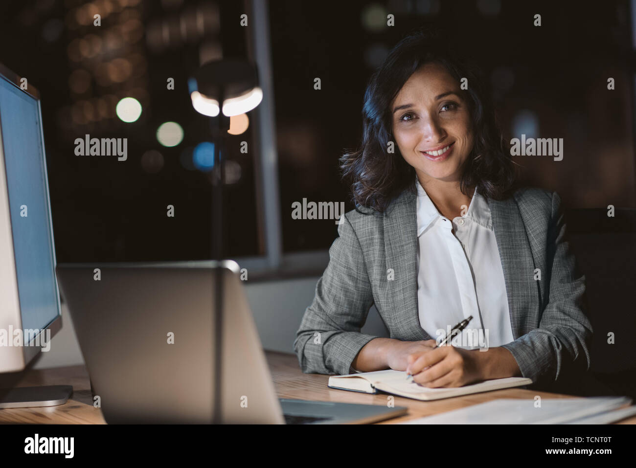 Ritratto di un sorridente giovane imprenditrice seduto alla sua scrivania in ufficio scrivere note e utilizzando un computer portatile mentre lavoro straordinario di notte Foto Stock