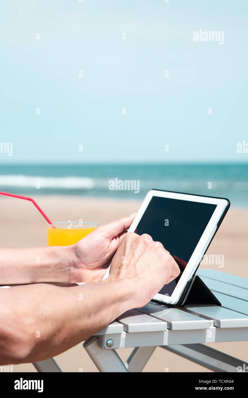 Primo piano di un giovane uomo caucasico sulla spiaggia, utilizzando una tavoletta digitale, seduti a un tavolo all'aperto, accanto a un bicchiere con una fresca bevanda a base di arancia, un Foto Stock