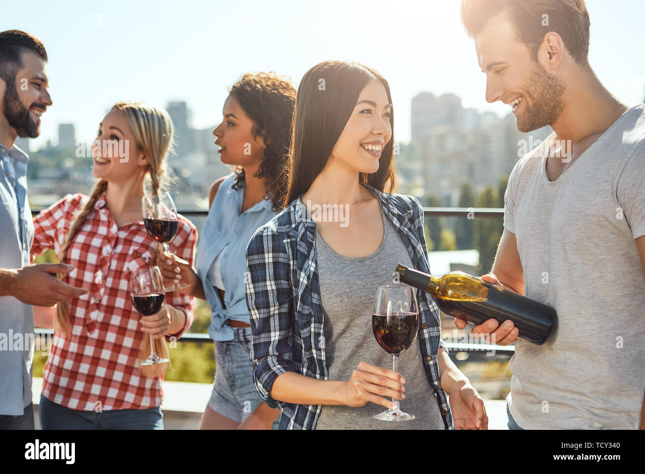 Divertirsi con gli amici. Giovane uomo barbuto è versando il vino per il suo amico e sorridente mentre si sta in piedi sul tetto con gli amici. Concetto di barbecue. Estate Foto Stock