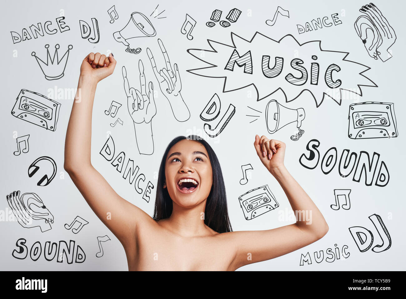 Dancing bellezza. Felice donna asiatica è il sollevamento in alto le mani e sorridenti mentre in piedi contro uno sfondo grigio con tema musicale scarabocchi su di esso. Musica e dan Foto Stock