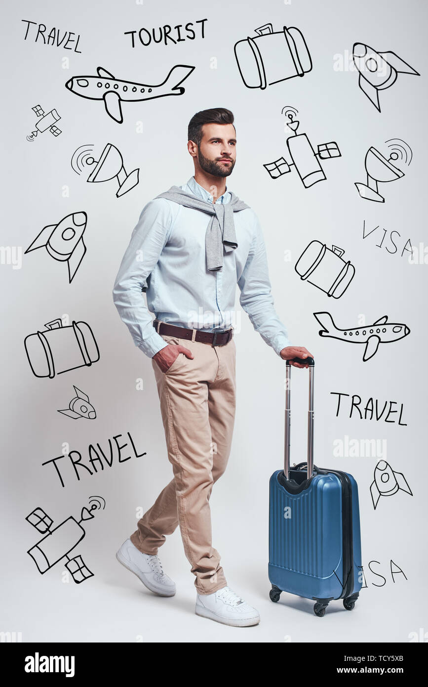 Concetto di viaggio. A lunghezza piena di fiducia e bello uomo barbuto trasportare il suo bagaglio mentre in piedi contro uno sfondo grigio con scarabocchi su di essi Foto Stock