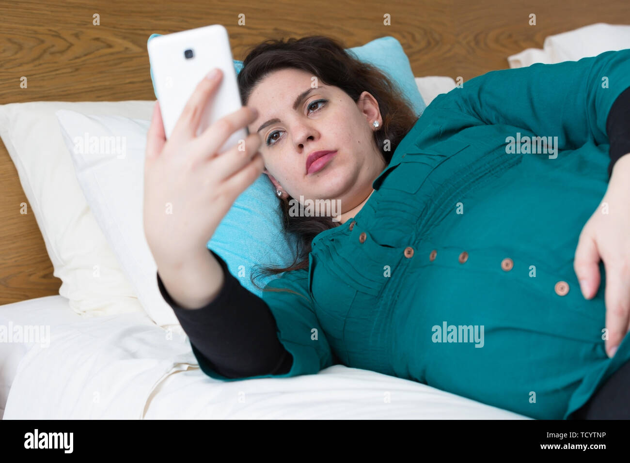 Donna incinta posa sul letto tramite il suo smartphone Foto Stock