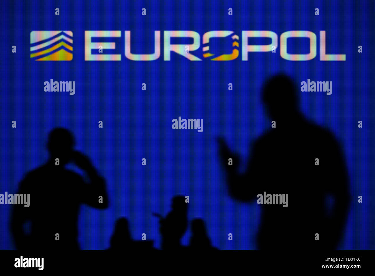 Il logo di Europol sia visibile su uno schermo a LED in background mentre si profila una persona utilizza uno smartphone in primo piano (solo uso editoriale) Foto Stock