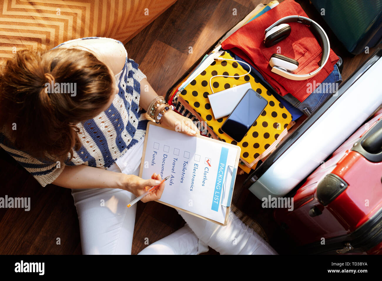 Vista superiore della donna elegante in bianco i pantaloni e camicia a righe con lista di imballaggio nei pressi di aprire la valigia di viaggio a casa moderna nella soleggiata giornata estiva. Foto Stock