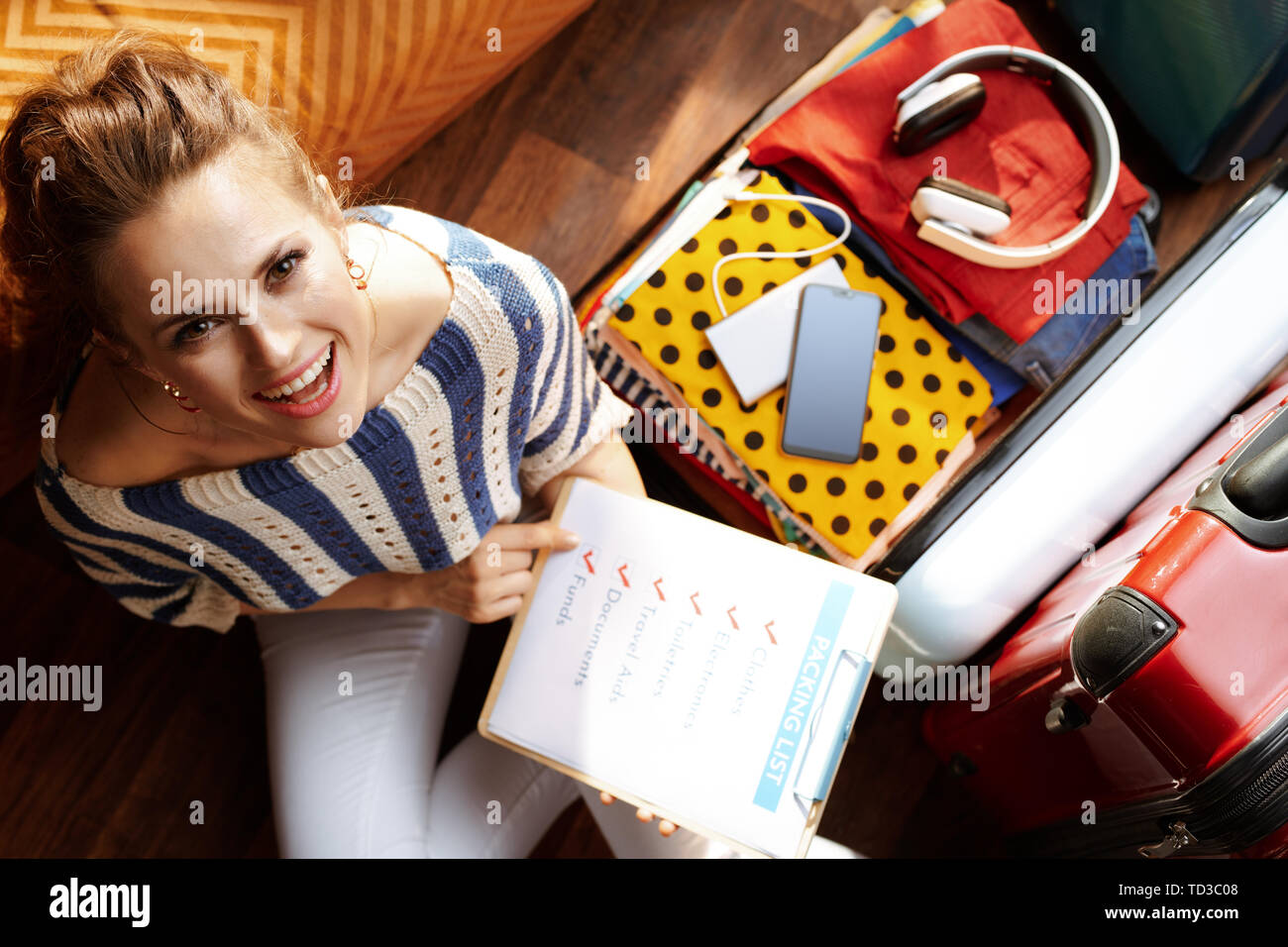 Vista superiore della sorridente giovane donna in bianco i pantaloni e camicia a righe con lista di imballaggio nei pressi di aprire la valigia di viaggio a casa moderna nella soleggiata giornata estiva. Foto Stock