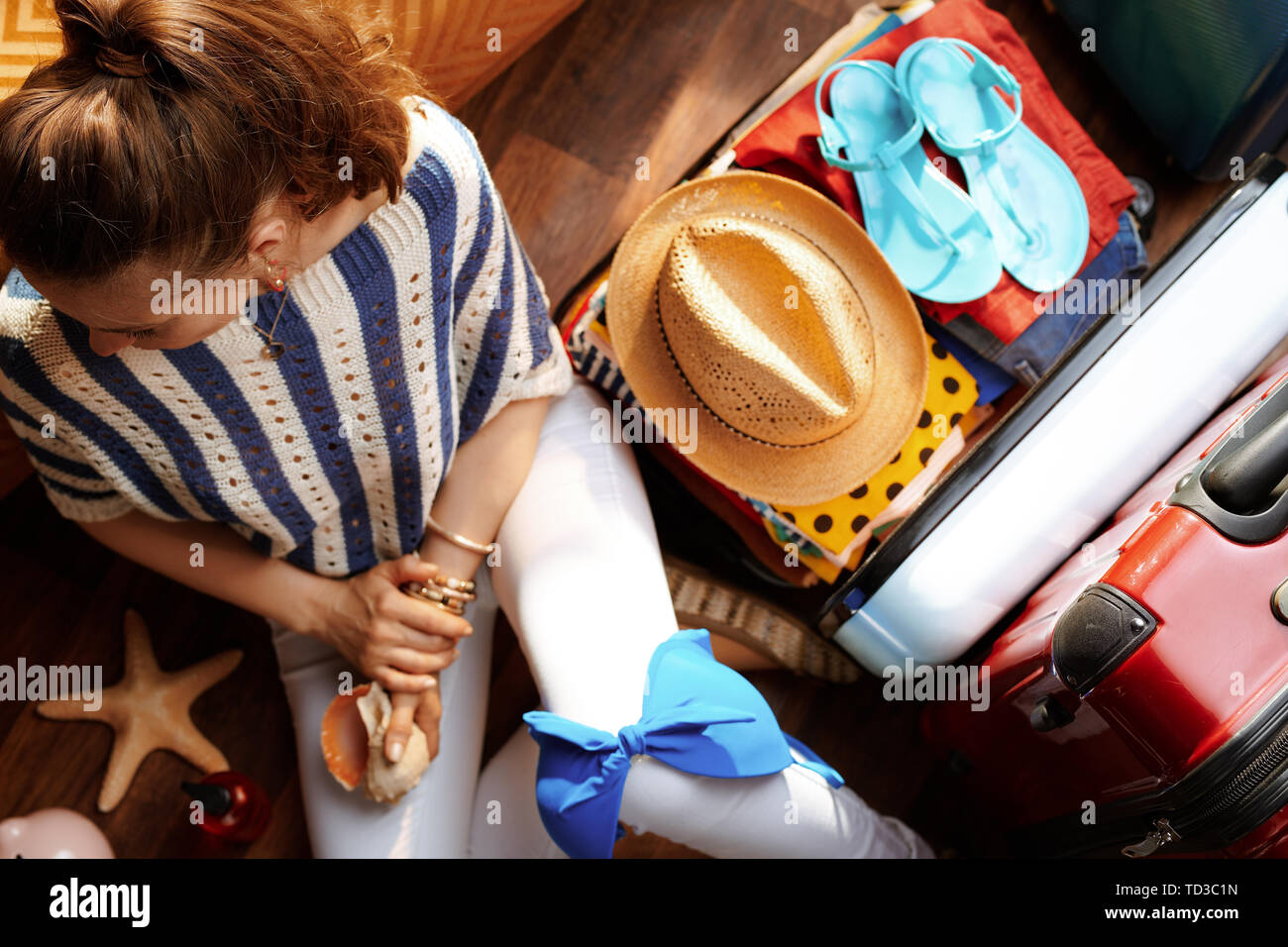 Vista superiore della donna elegante in bianco i pantaloni e camicia a righe nei pressi di aprire la valigia di viaggio con vacanze estate roba in soggiorno moderno nella soleggiata s Foto Stock