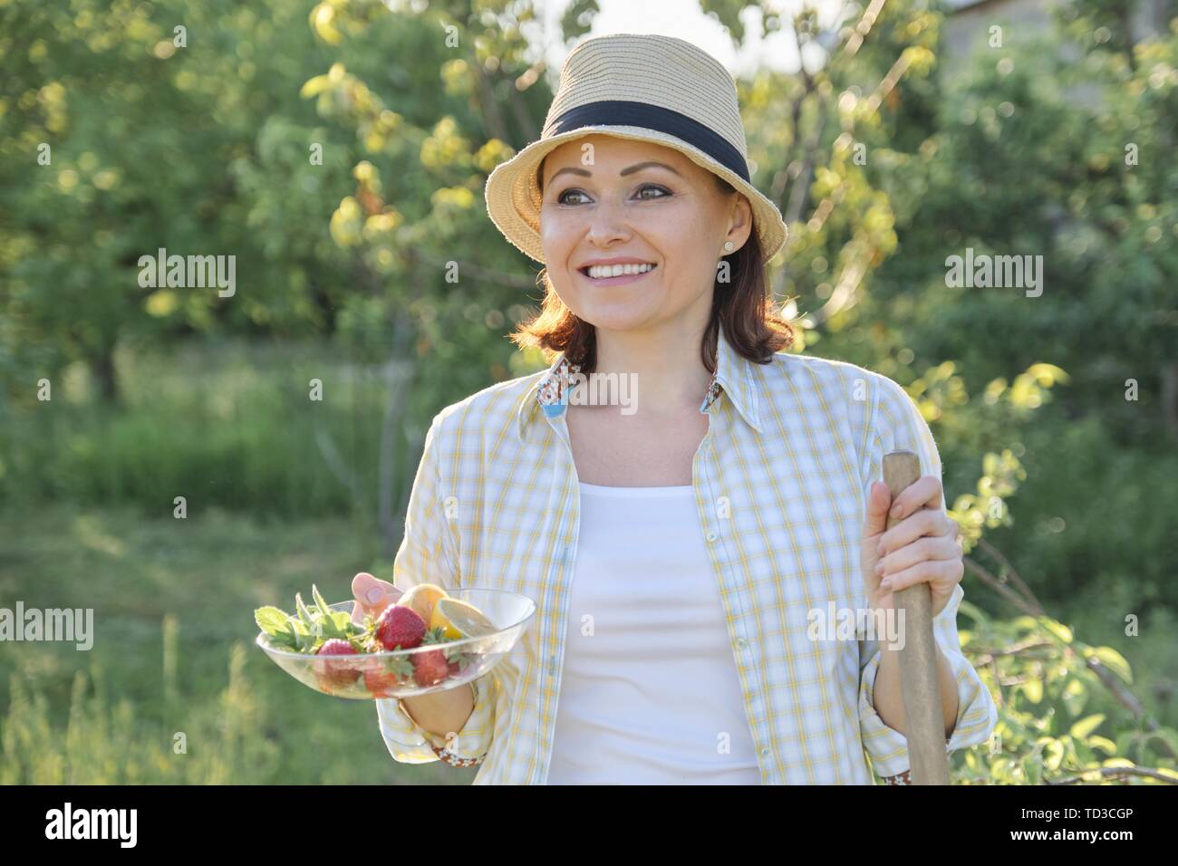 Outdoor ritratto di donna felice 40 anni, femmina in giardino nel cappello di paglia con la piastra di fragole limone menta, estate vacanze e lavoro in natura Foto Stock