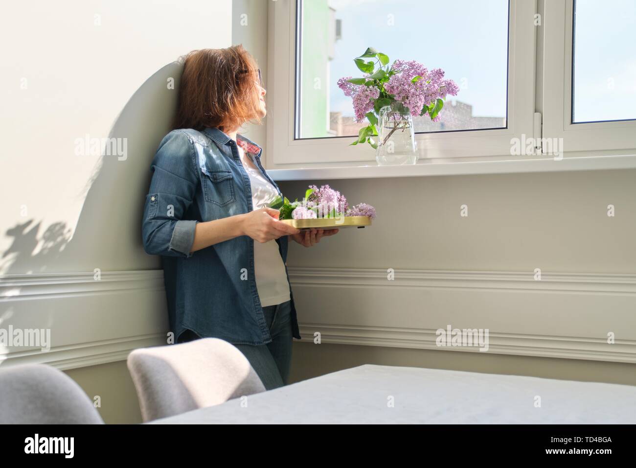 Ritratto di bella donna di mezza età in casa con bouquet di fiori lilla. Sullo sfondo la parete di luce solare nella finestra spazio di copia Foto Stock