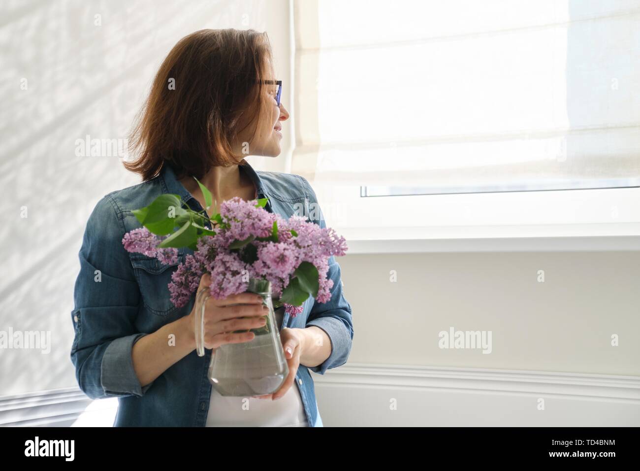 Ritratto di bella donna di mezza età in casa con bouquet di fiori lilla. Sullo sfondo la parete di luce solare nella finestra spazio di copia Foto Stock