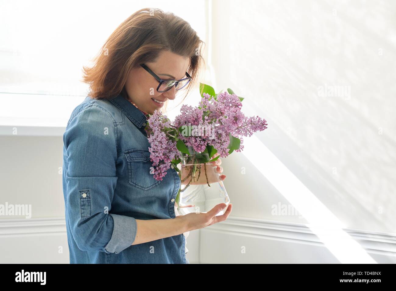 Ritratto di bella donna di mezza età in casa con bouquet di fiori lilla. Sullo sfondo la parete di luce solare nella finestra spazio di copia Foto Stock