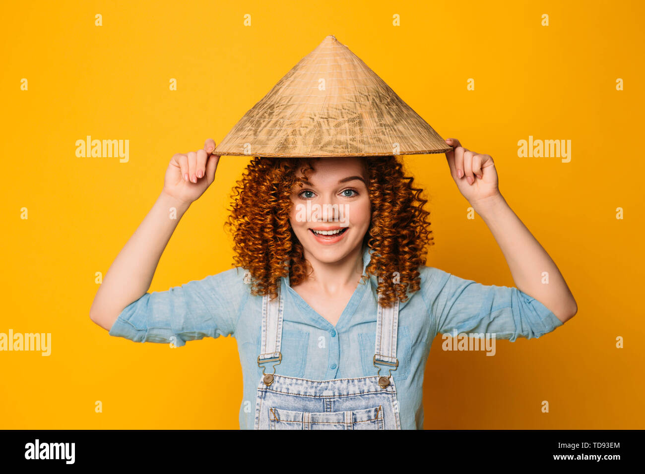 I capelli rossi ricci donna in un cappello cinese è ridere, su uno sfondo giallo. Viaggio in Oriente, Cina e Asia. Foto Stock