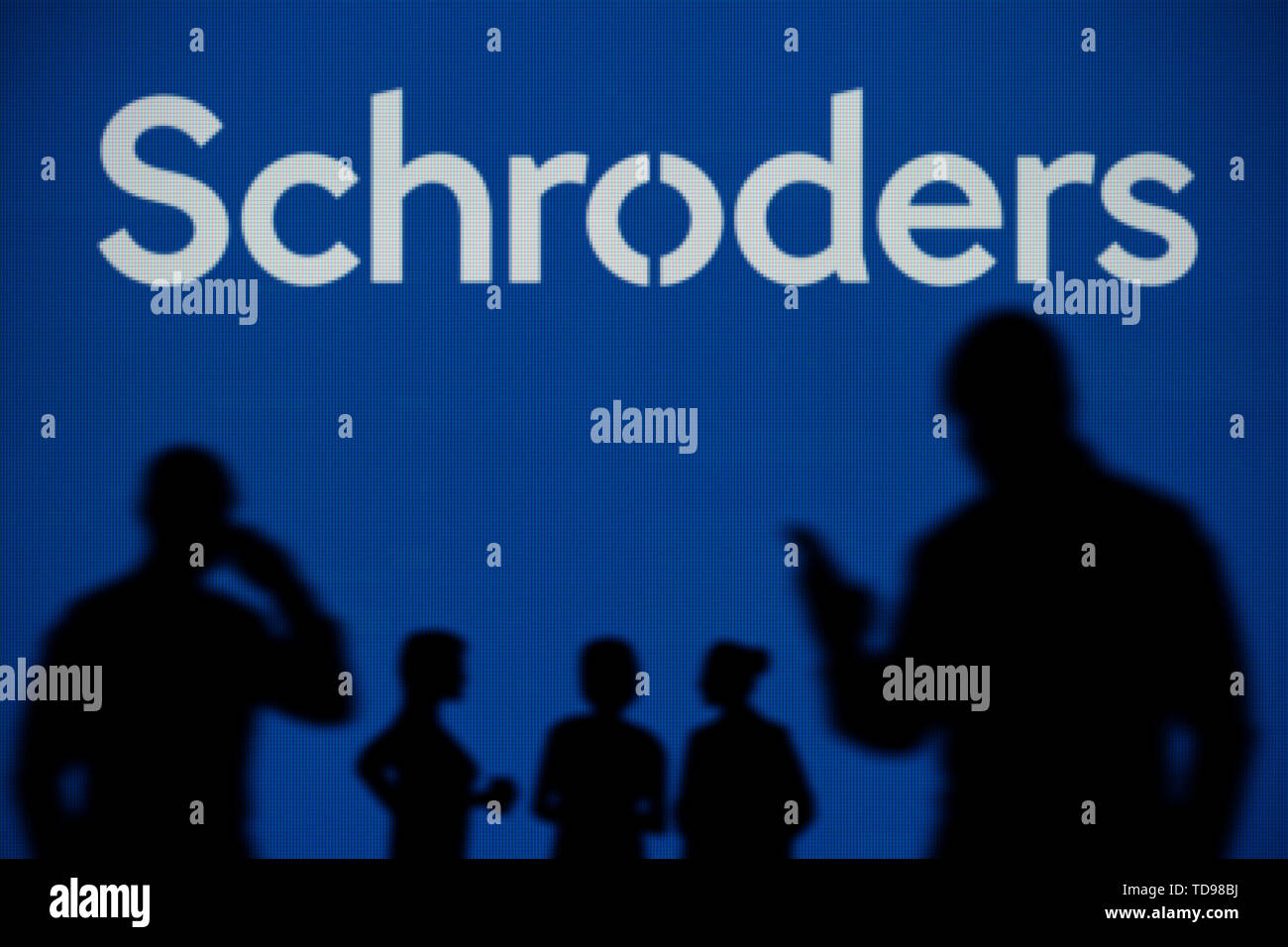 La Schroders logo è visibile su uno schermo a LED in background mentre si profila una persona utilizza uno smartphone in primo piano (solo uso editoriale) Foto Stock
