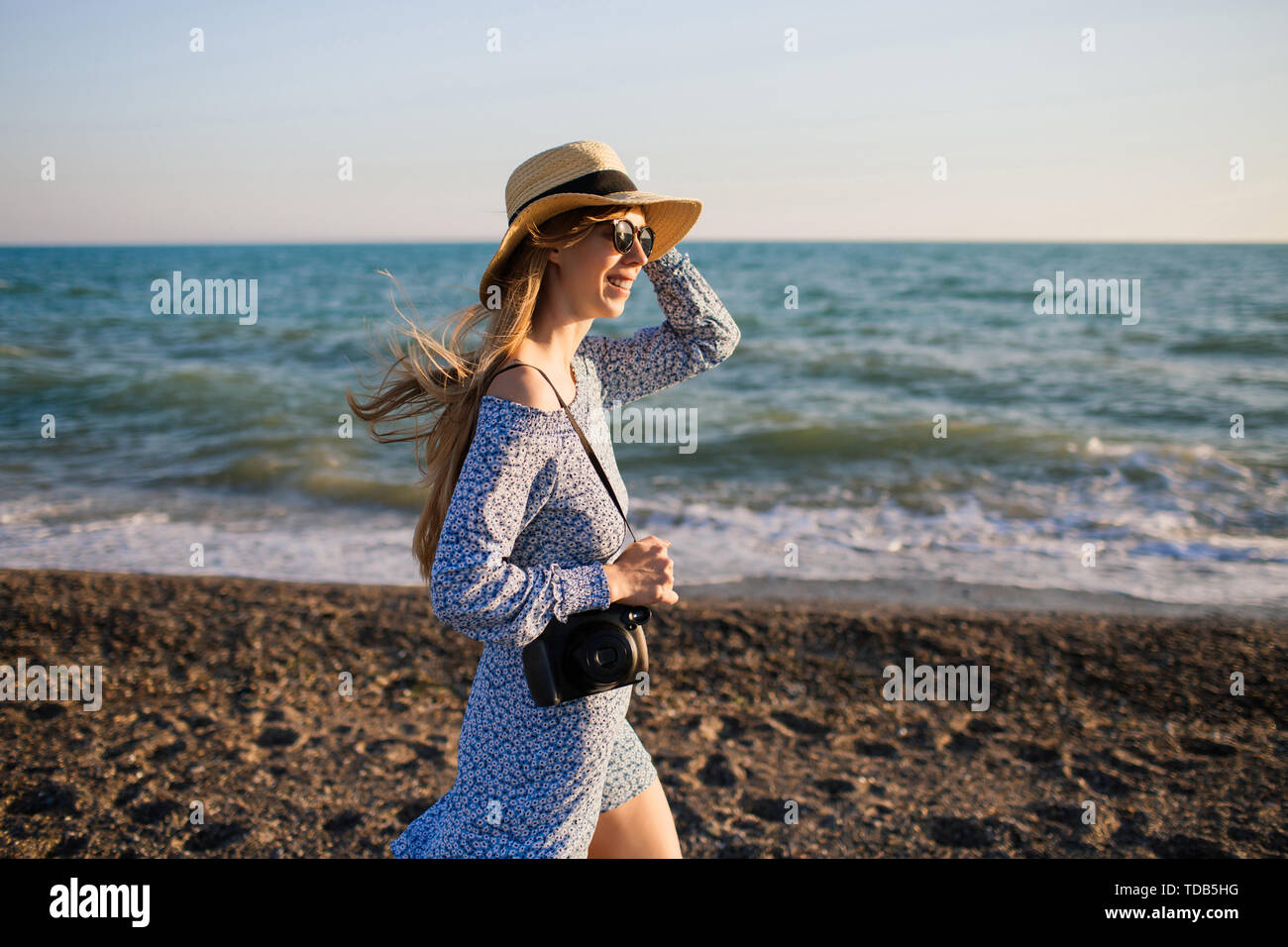 Giovane attraente ragazza sorridente con lunghi capelli biondi, indossando un cappello, passeggiate lungo la spiaggia. Azienda fotocamera nero. Onde blu sullo sfondo. Foto Stock