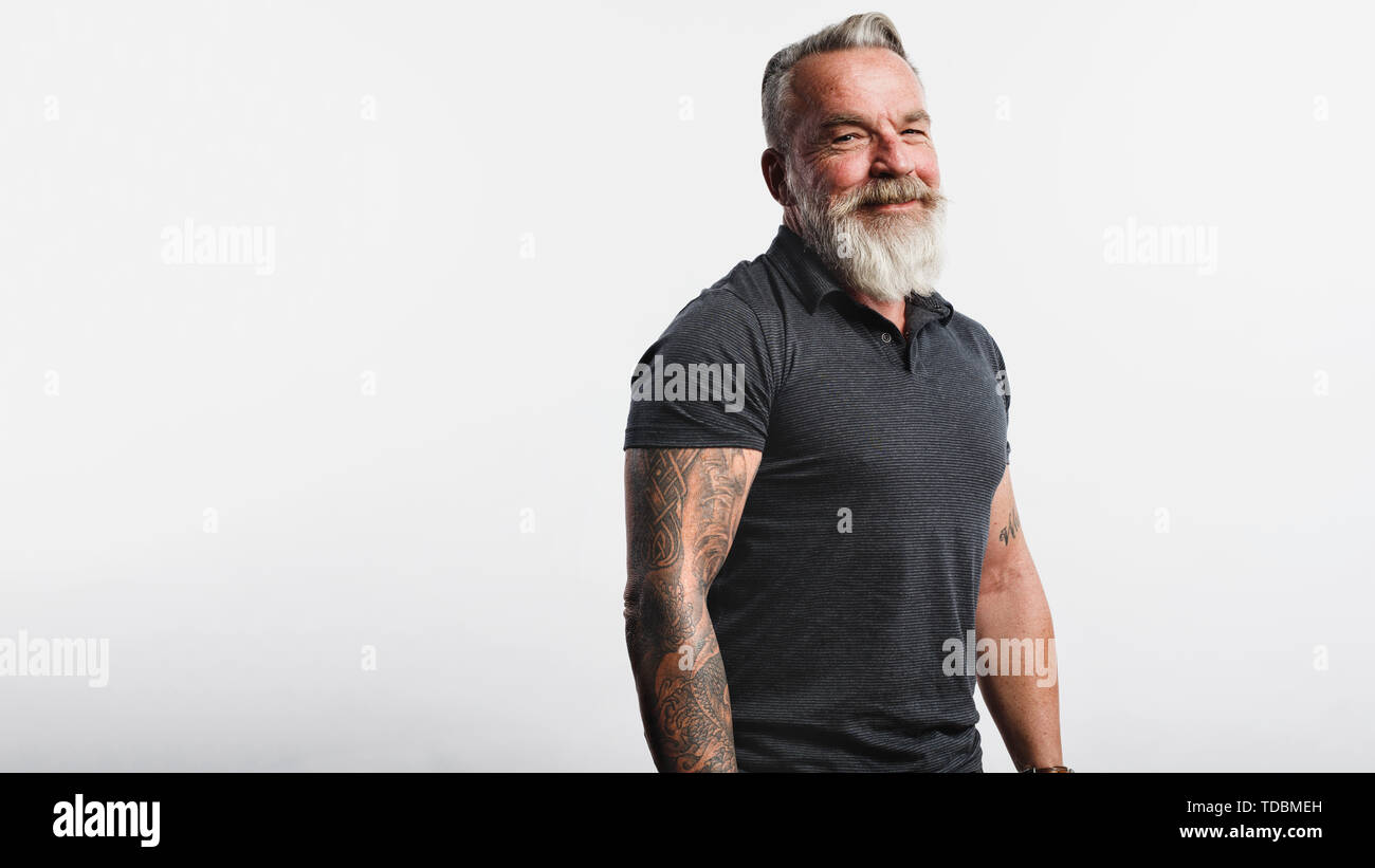 Ritratto di vecchio maschio muscolare con la barba bianca guardando la fotocamera. Senior sorridente uomo con tatuaggio su bracci in piedi contro uno sfondo bianco. Foto Stock