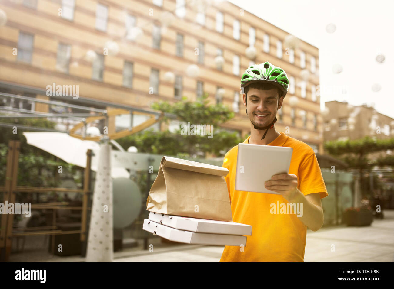 Giovane uomo in maglietta gialla consegna pizza con i gadget per via ordine presso la città della strada. Corriere on-line utilizzando app per ricevere il pagamento e il tracking spedizioni indirizzo. Le moderne tecnologie. Foto Stock