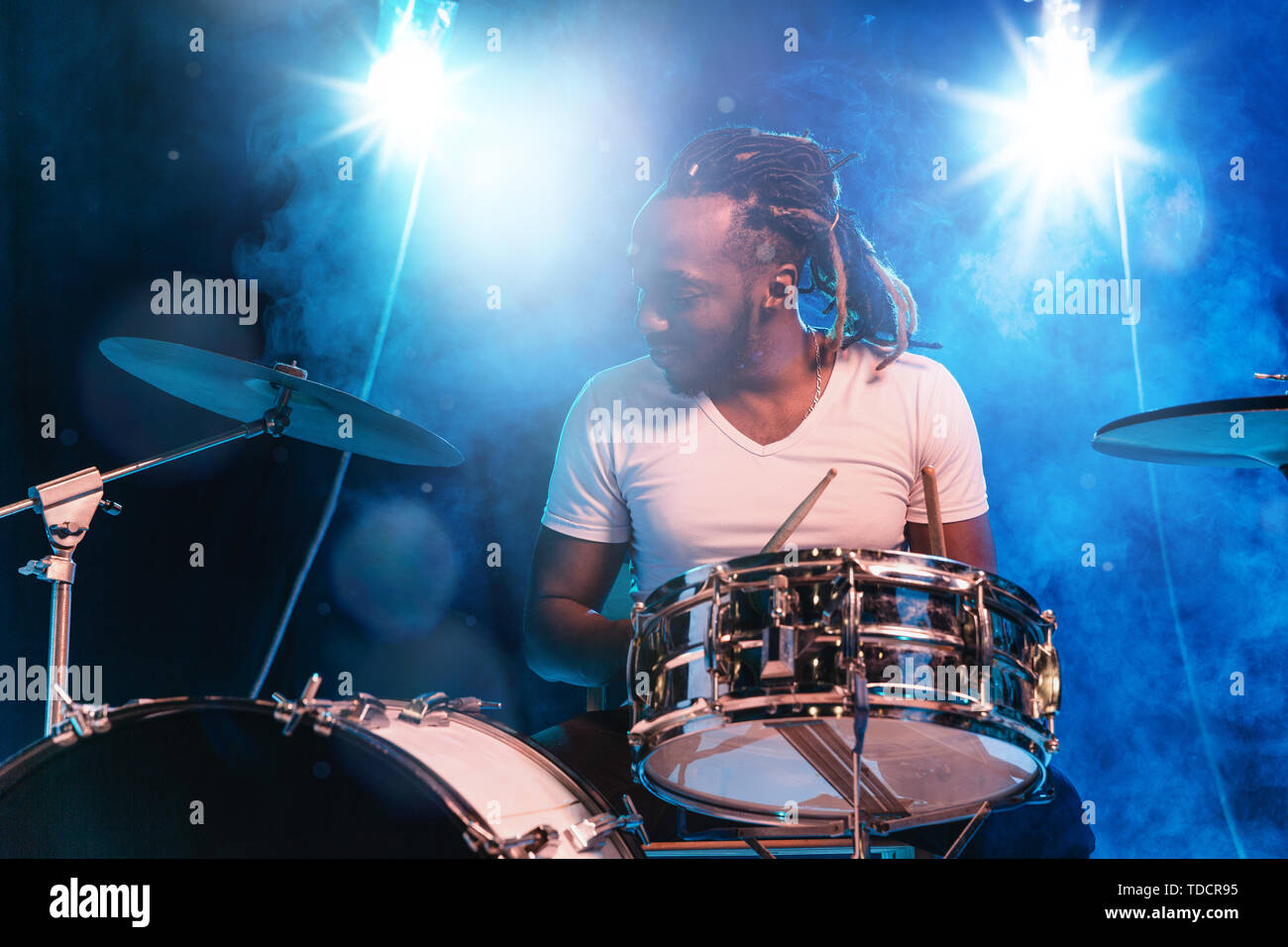 Giovani afro-americani musicista jazz o il batterista suonando la batteria  su blu di sfondo per studio in fumo incandescente attorno a lui. Concetto  di musica, hobby, inspirness. Ritratto colorato di gioiosa artista