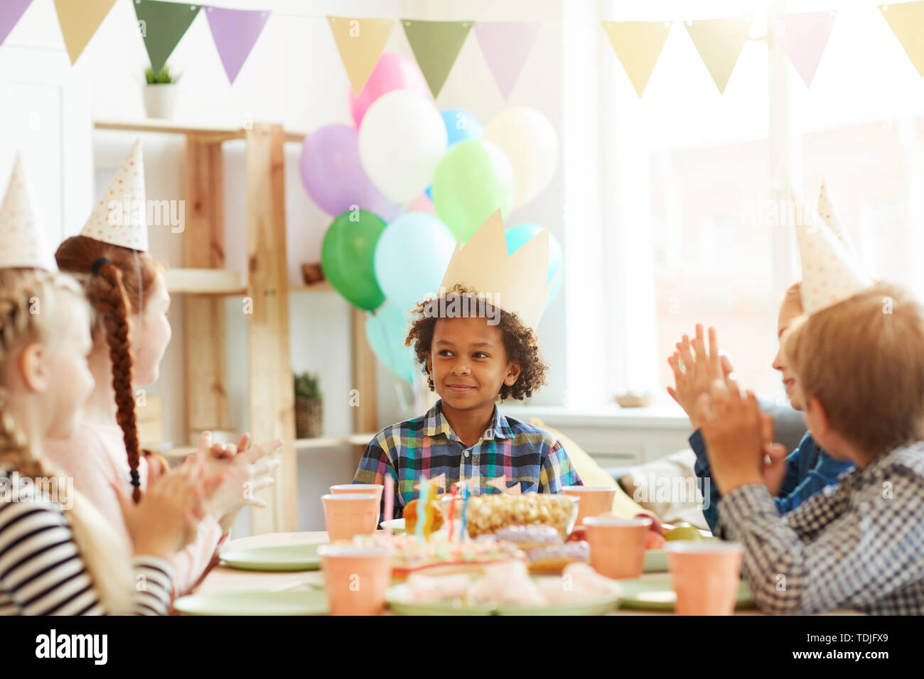 Ritratto di sorridente ragazzo afro-americano che indossa la corona seduta a tavola mentre festeggia il compleanno con gli amici, spazio di copia Foto Stock